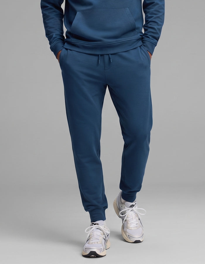 Model is wearing the Cozy Earth Men's Versa Hoodie & Jogger Set in blue, paired with white athletic sneakers, standing with hands in pockets against a plain gray background.|Color:Obsidian
