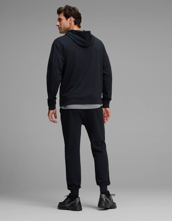A man stands facing away, wearing the Cozy Earth Men's Versa Hoodie & Jogger Set in black. He is standing on a light gray floor with a plain gray background.|Color:Jet Black