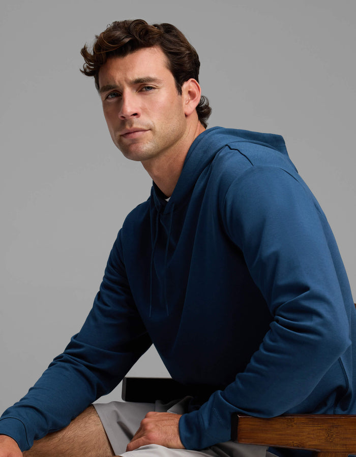 A man with short brown hair wears the Men's Versa Hoodie by Cozy Earth in blue and light gray shorts as he sits on a wooden chair, gazing slightly to the side with a neutral expression against a plain gray background.|Color:Obsidian