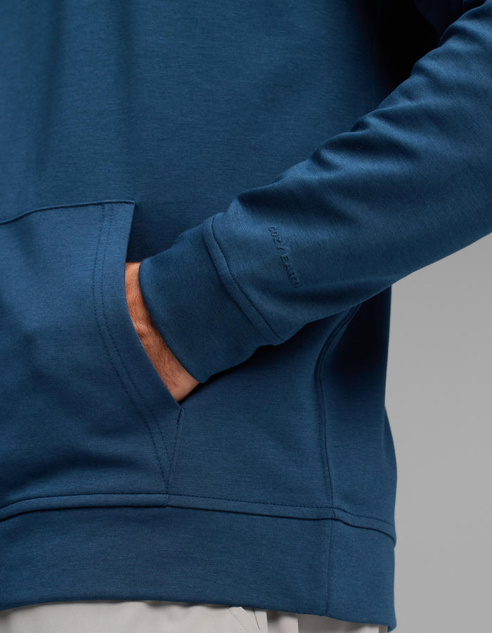 A man wearing the Cozy Earth Men's Versa Hoodie in blue, with one hand in the front pocket, stands against a plain gray background.|Color:Obsidian
