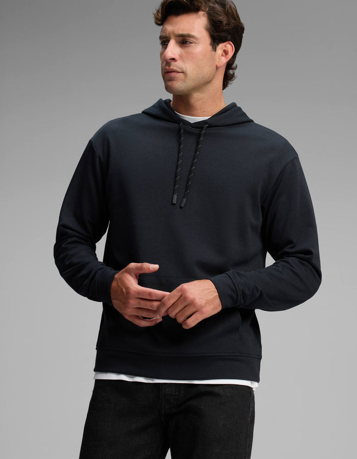 A man wearing the Cozy Earth Men's Versa Hoodie and dark jeans stands against a plain gray background, looking to his left with his hands loosely clasped in front of him.|Color:Jet Black