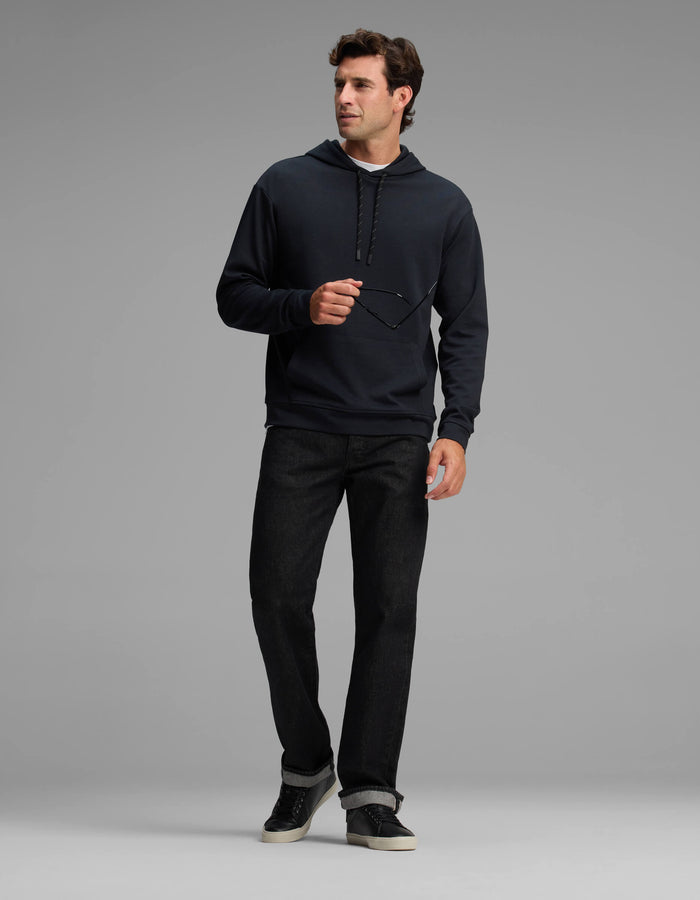 A man in the Cozy Earth Men's Versa Hoodie and black jeans stands against a plain gray background, holding eyeglasses in one hand and looking to the side.|Color:Jet Black