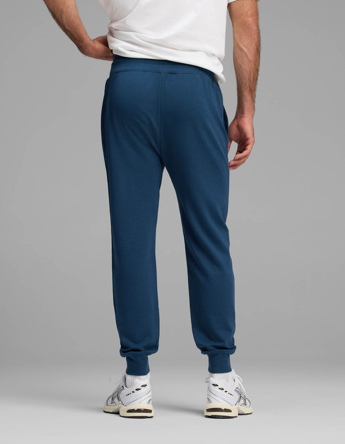 A person is standing with their back to the camera against a plain gray background, wearing a white T-shirt, white sneakers, and blue Cozy Earth Men's Versa Jogger pants.|Color:Obsidian