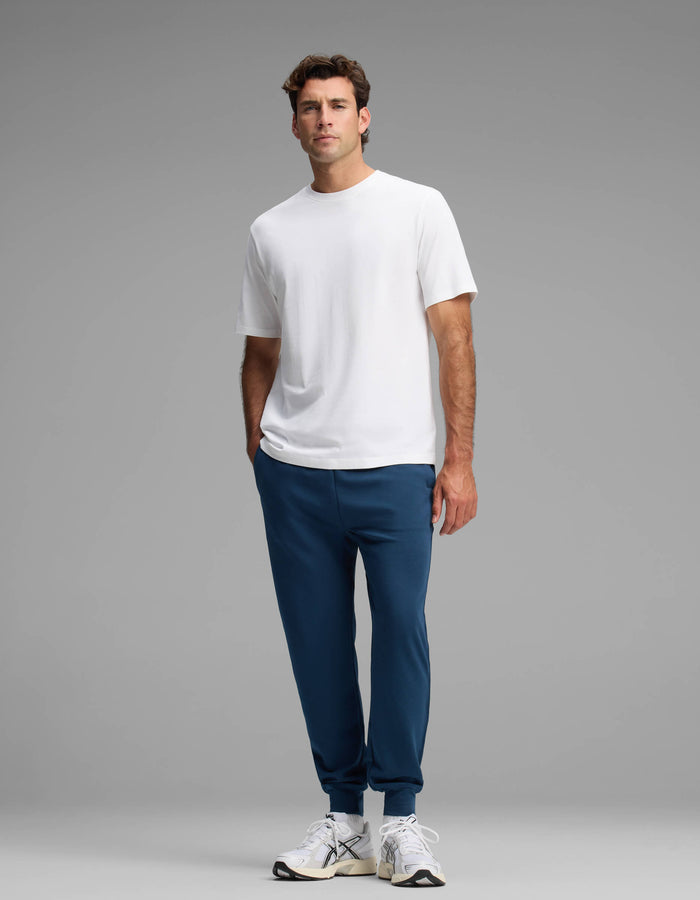 A man stands against a gray background wearing a plain white t-shirt, white sneakers, and Cozy Earth's Men's Versa Jogger in blue, with his left hand in his pocket.|Color:Obsidian