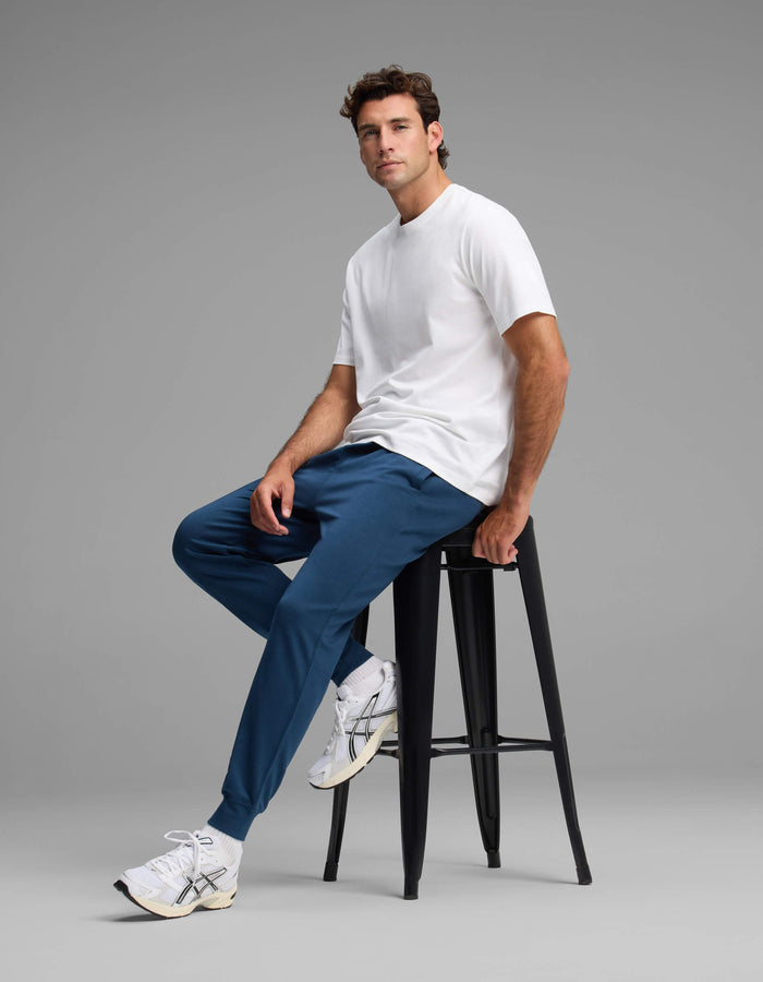 A man wearing the Cozy Earth Men's Versa Jogger in blue and a white t-shirt sits on a tall black stool against a plain gray background, paired with white sneakers and gazing slightly to the side.|Color:Obsidian