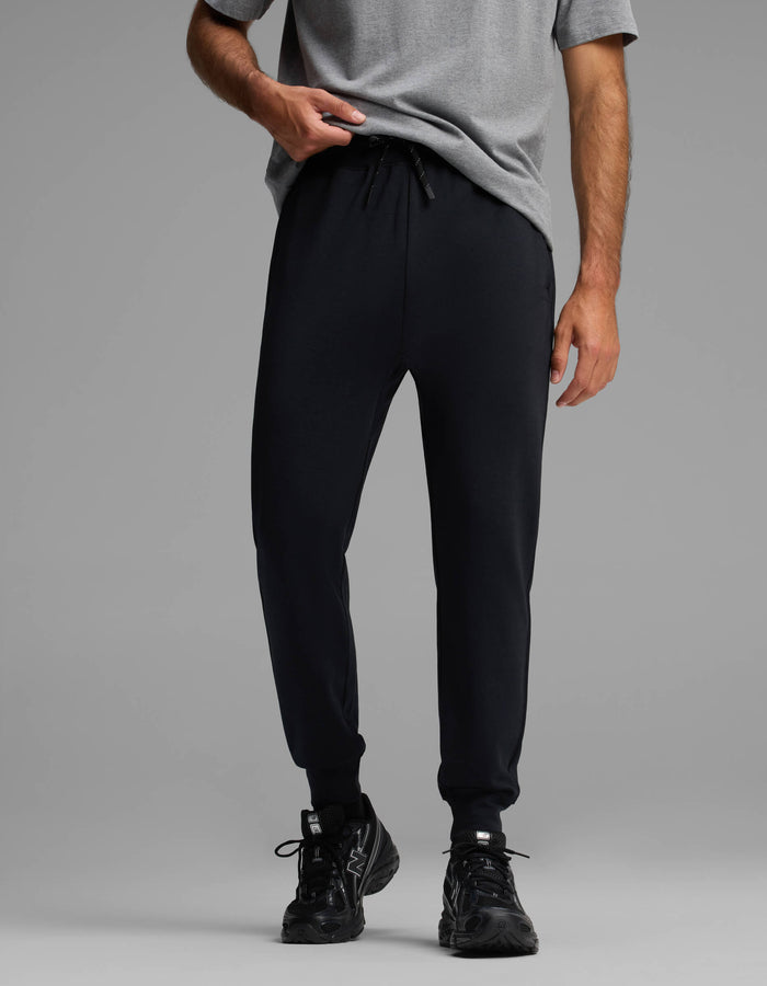A person wearing Cozy Earth's Men's Versa Jogger in black and a grey T-shirt stands against a plain grey background, holding the hem of their shirt, with only their lower torso and legs visible.|Color:Jet Black