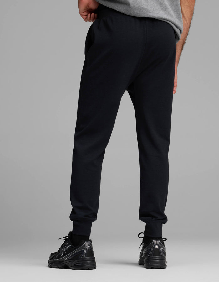 A person stands facing away, wearing a gray T-shirt, Cozy Earth Men's Versa Jogger sweatpants in black, and black sneakers against a plain gray background.|Color:Jet Black