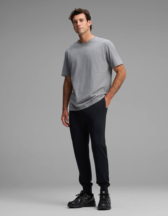 A man stands against a plain gray background, wearing a gray short-sleeve t-shirt, black sneakers, and the Cozy Earth Men's Versa Jogger in black, with one hand in his pocket.|Color:Jet Black