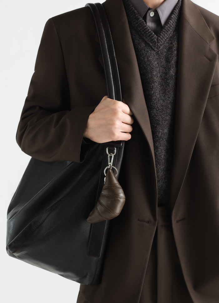 Mini croissant leather keyring in dark brown attached to black leather tote worn by model LEMAIRE