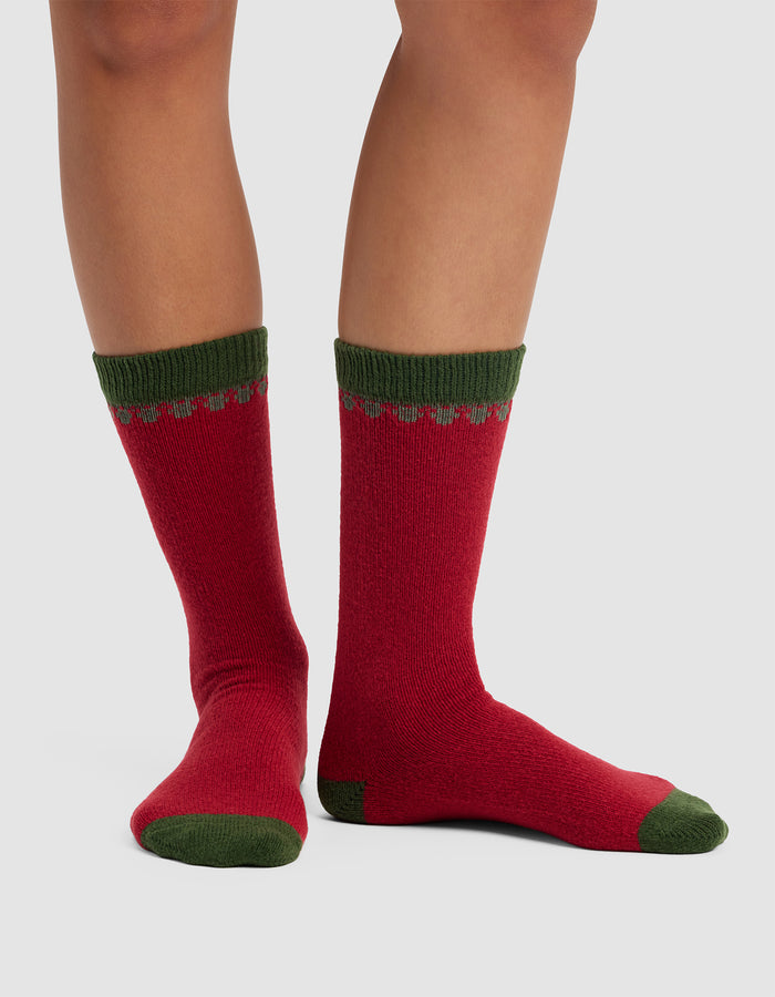 A pair of legs wear Cozy Earth's The Mistletoe Plush Lounge Socks, featuring red with green heels, toes, and cuffs plus a scalloped green and red design near the top. Set against a plain light gray background.|Color:Cranberry