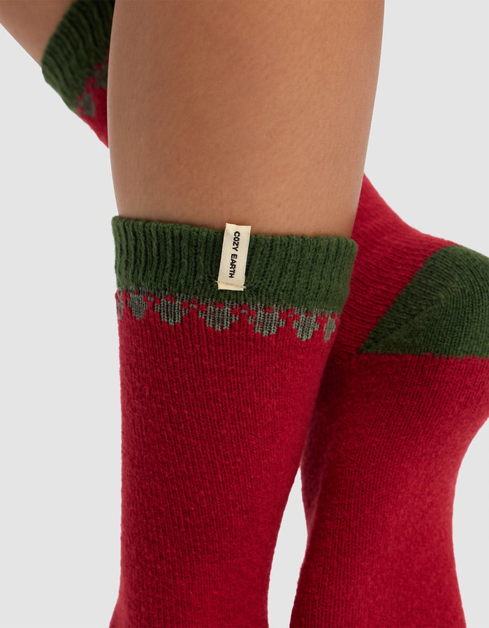 Close-up of a person's legs wearing The Mistletoe Plush Lounge Sock by Cozy Earth—cozy red socks with green cuffs, a small beige label, and a decorative pattern near the top.|Color:Cranberry