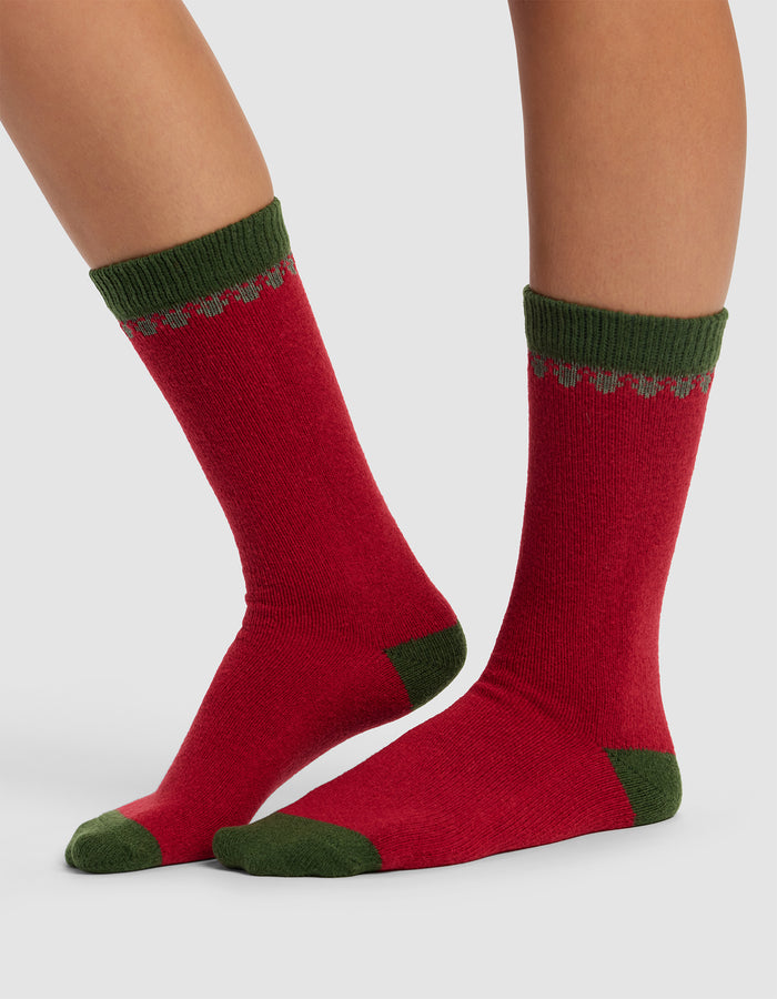 A person wears The Mistletoe Plush Lounge Sock by Cozy Earth, featuring red with green heels, toes, cuffs, and a green decorative top pattern, standing on a light gray background.|Color:Cranberry