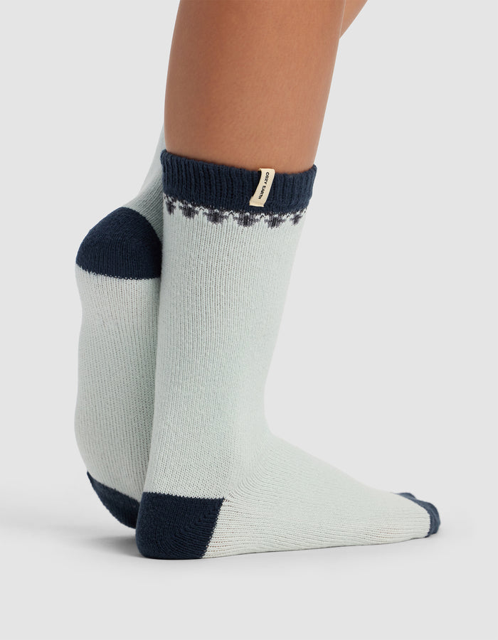 A person wears The Mistletoe Plush Lounge Sock by Cozy Earth—light gray crew socks with navy blue toes, heels, and cuffs, a small decorative pattern near the cuff, and a beige tag. Their feet are crossed at the ankles.|Color:Mist