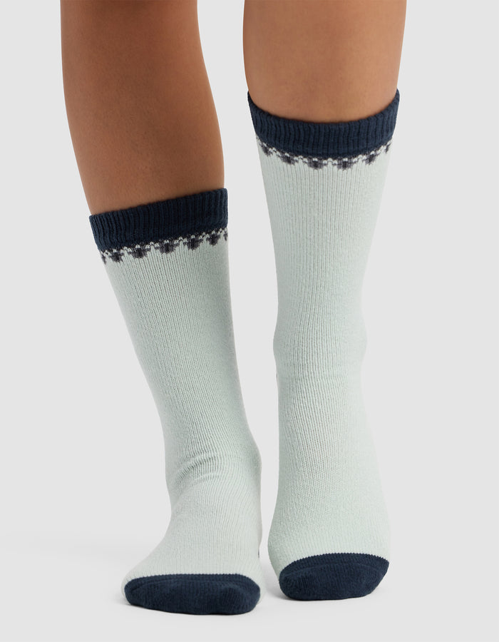 Someone wearing The Mistletoe Plush Lounge Sock by Cozy Earth, featuring light fabric with dark blue toes, heels, and cuffs, plus a decorative blue pattern at the top, stands on a light surface.|Color:Mist