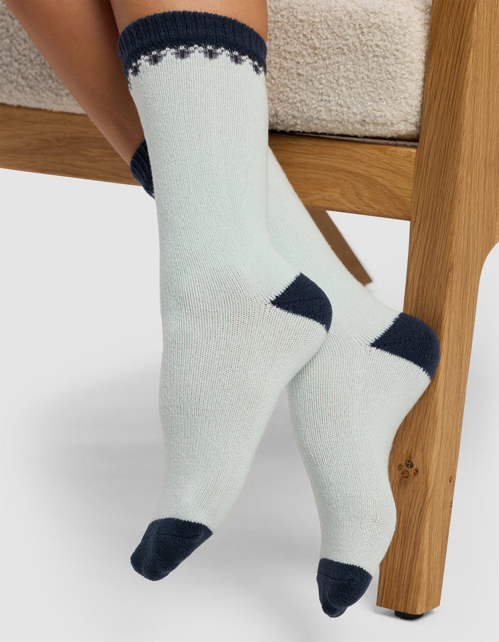 Close-up of a person wearing Cozy Earth's The Mistletoe Plush Lounge Sock, featuring light fabric with dark heels, toes, and cuffs, while sitting on a wooden chair with crossed feet on a neutral background.|Color:Mist