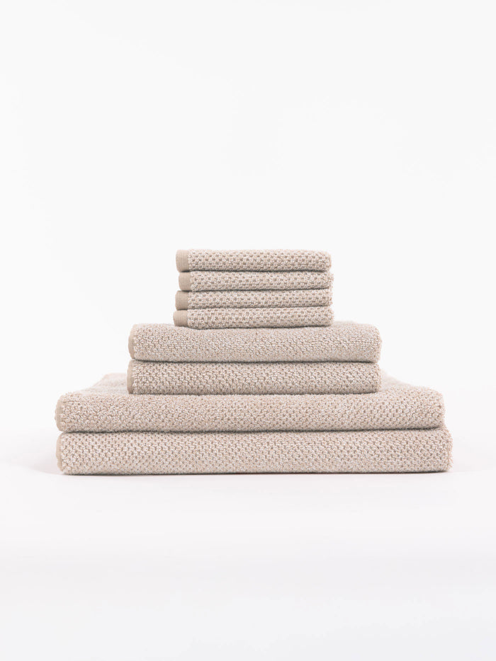 The Cozy Earth Nantucket Bath Towel Set features beige textured towels in various sizes, neatly stacked from largest to smallest, set against a plain white background.|Color:Heathered Sand