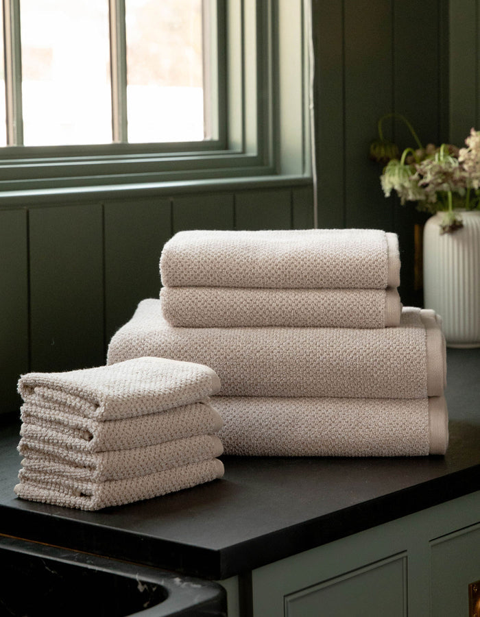 The Cozy Earth Nantucket Bath Towel Set, featuring hand and bath towels in beige, is neatly stacked on a dark countertop near a window with green paneling. A white vase with flowers appears in the background.|Color:Heathered Sand