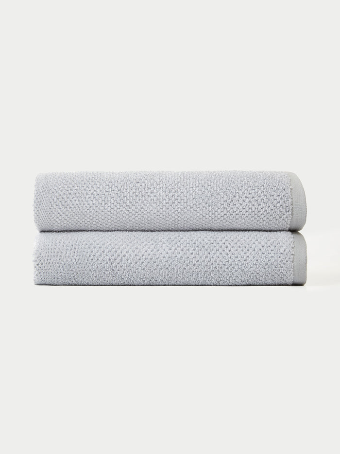The Nantucket Bath Towel Set by Cozy Earth features two neatly folded light gray towels stacked together against a plain white background.|Color: Heathered Harbor Mist