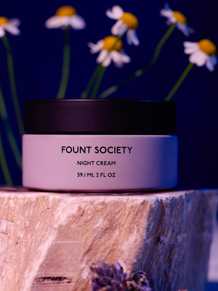 A jar of Cozy Earth Night Cream sits on a wooden surface, its label clearly visible. In the backdrop, soft-focus white daisies with yellow centers stand out against a blue background. |Size:Full Size