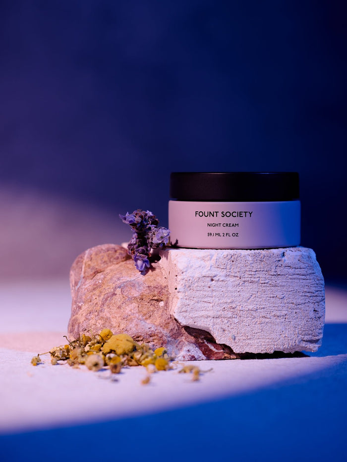 A jar of Cozy Earth Night Cream rests on a textured stone, surrounded by small dried flowers under gentle purple and pink lighting. |Size:Full Size