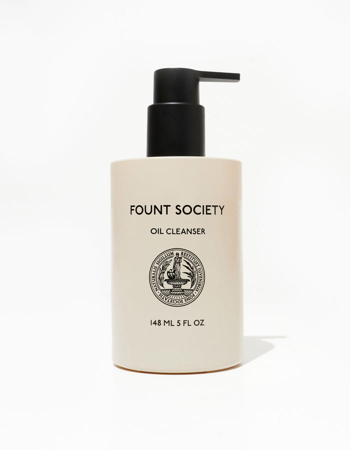 A pump bottle of "Cozy Earth Oil Cleanser" is shown. The bottle is a sleek, ivory-colored container with a black pump dispenser. The minimalist label on the front contains the brand name, product name, and volume of 148 ml (5 fl oz) along with a decorative logo.