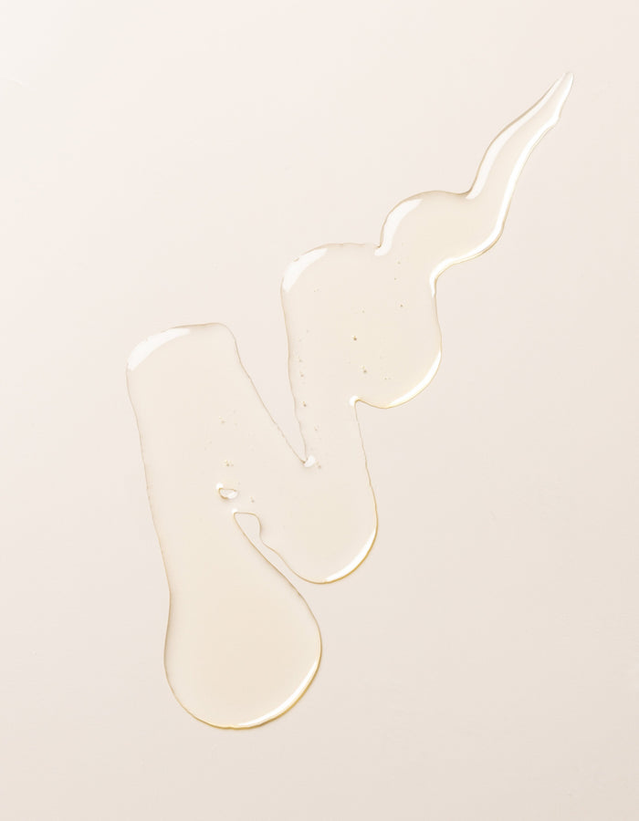 An irregular-shaped spill of Cozy Earth's Oil Cleanser on a smooth, light-colored surface. The clear liquid forms a long, winding pattern with droplets and uneven edges, creating the impression of fluid motion.