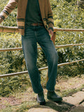 A man outdoors wearing blue denim jeans, a brown jacket, a dark green shirt, and dark rain boots, standing near a bamboo fence with green foliage in the background.