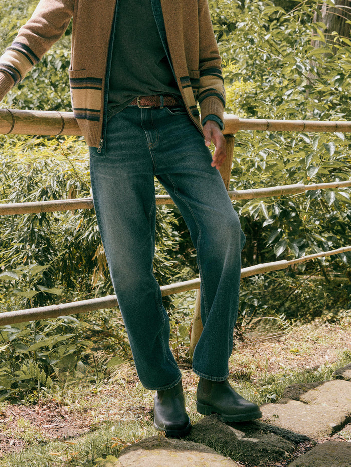 A man outdoors wearing blue denim jeans, a brown jacket, a dark green shirt, and dark rain boots, standing near a bamboo fence with green foliage in the background.