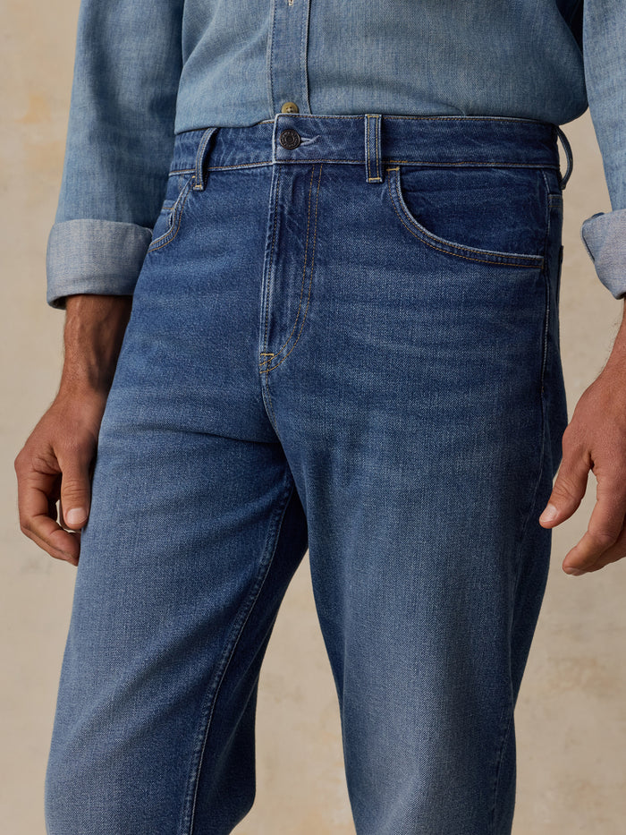 Close-up of a person wearing medium blue denim jeans with a rolled-up light blue denim shirt, shown from the waist to upper thighs against a warm beige textured background.