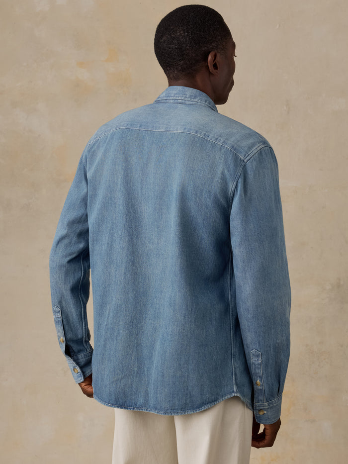A man viewed from the back wearing a light blue denim long-sleeve button-up shirt and matching blue jeans, standing against a plain beige wall.