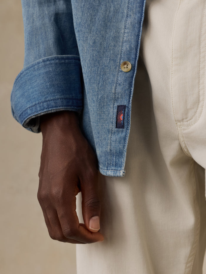Close-up of a light blue denim button-up shirt showing the button closure, fabric texture, and a small Faherty brand sun and waves patch near the hem