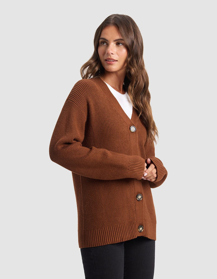 A woman with long brown hair wears the Cozy Earth Oversized Classic Cardigan, featuring a ribbed texture and large buttons, layered over a white top and black pants against a plain light gray background.|Color:Spice