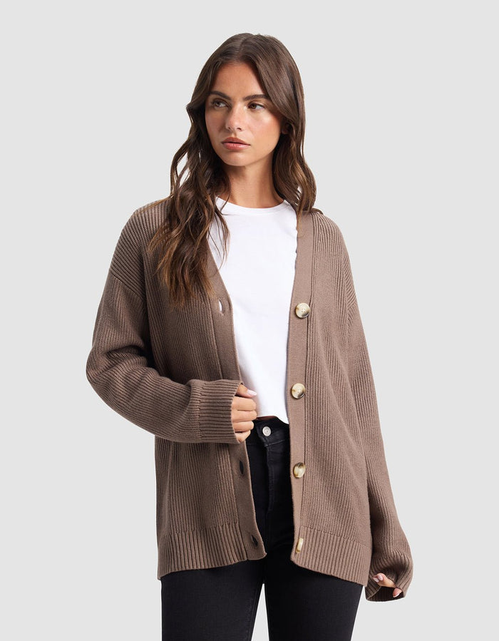 A woman stands against a light gray background, wearing Cozy Earth’s Oversized Classic Cardigan in brown with large buttons, layered over a white t-shirt and black pants.|Color:Ebony