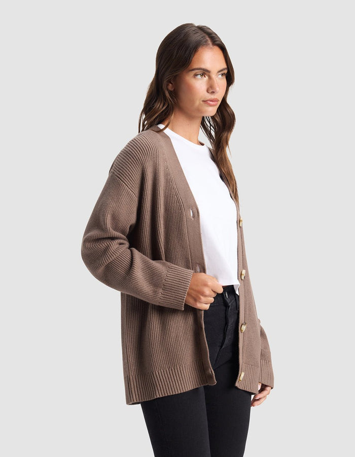 A woman stands against a light gray background, wearing the Cozy Earth Oversized Classic Cardigan in taupe over a white t-shirt and black pants, looking slightly to the side.|Color:Ebony