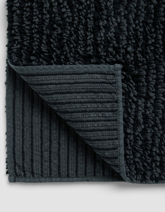 Close-up of the Cozy Earth Plush Loop Bath Mat with a corner folded, highlighting its ribbed, non-slip underside and thick, soft plush pile on top.|Color:Graphite