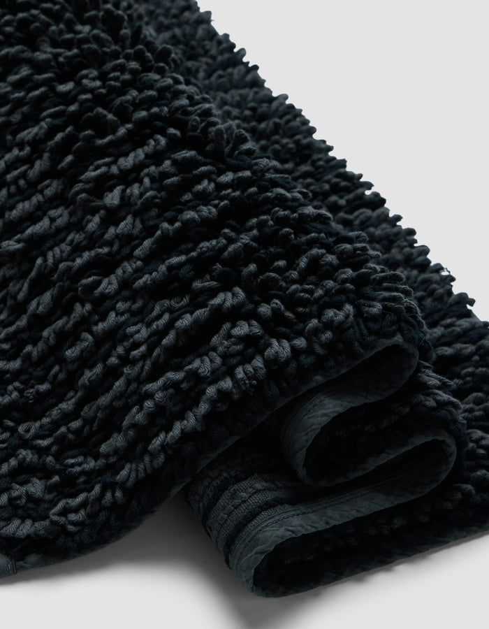A close-up of the Plush Loop Bath Mat by Cozy Earth shows its dark, textured fabric with a thick, looped pile and soft, plush folds against a plain light background.|Color:Graphite