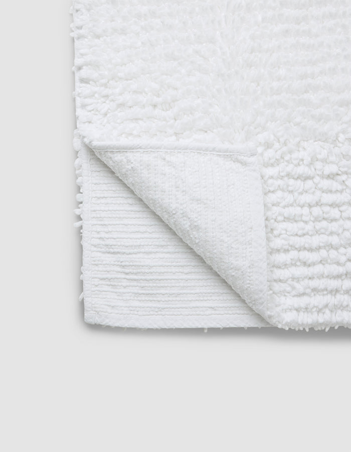 A close-up of the Cozy Earth Plush Loop Bath Mat in white, featuring a soft, textured surface with one corner flipped to show its flat underside. The mat is set against a plain light gray background.|Color:White