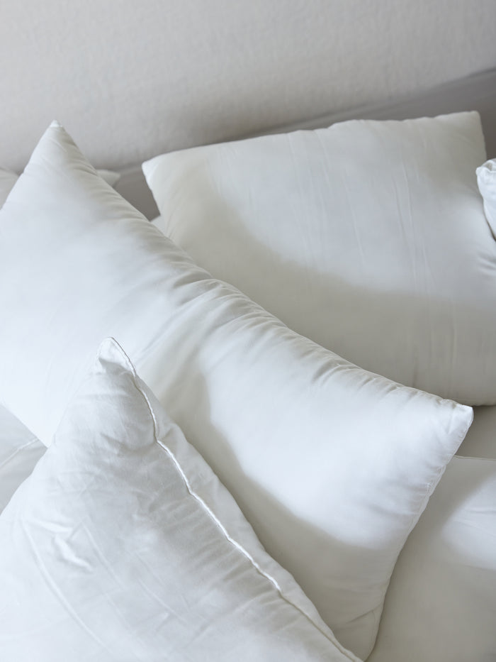 A close-up view of several **Cozy Earth Premium Down Alternative Inserts** arranged on a bed with white sheets. The pillows, varying slightly in shape and size, create a soft and cozy atmosphere. The background features a muted light color, enhancing the serene and clean setting. |Size:14" x 30"