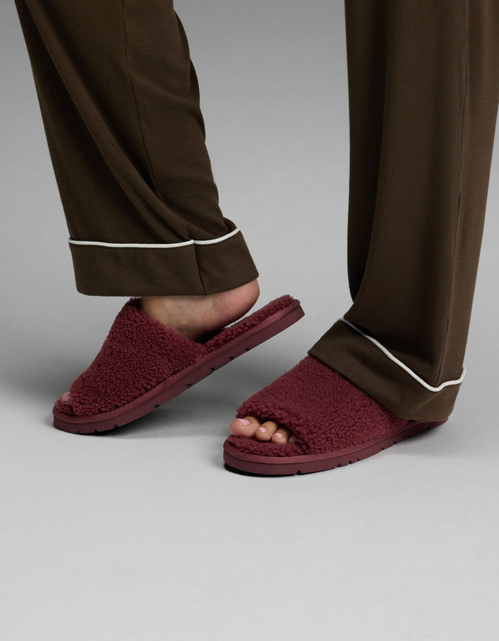 A person is wearing Cozy Earth’s brown pajama pants with white trim and Puffy Sheep Slides—maroon fuzzy open-toe slippers—while standing on a gray surface.|Color:Mulberry