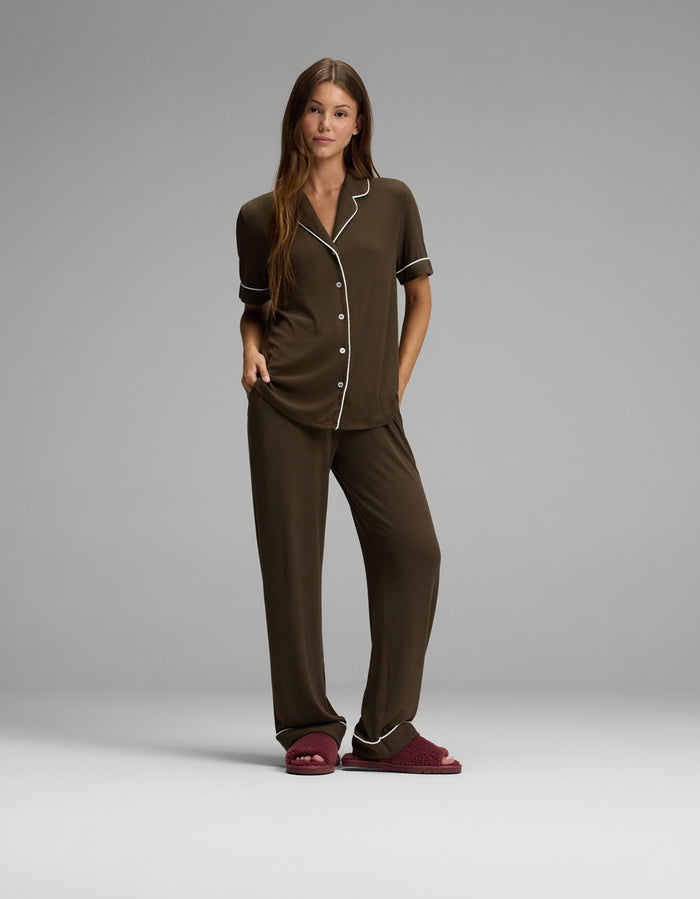 A woman stands wearing brown pajamas with white piping and Cozy Earth Puffy Sheep Slides. She has long brown hair and a relaxed posture against a plain gray background.|Color:Mulberry