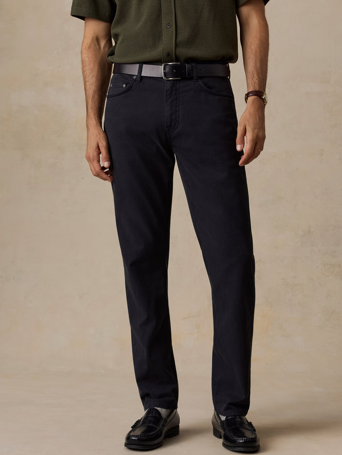 A man wearing dark navy blue slim-fit trousers, styled with an olive green shirt, a black belt, and black leather loafers, standing against a textured beige studio backdrop.