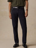 A man wearing dark navy blue slim-fit trousers, styled with an olive green shirt, a black belt, and black leather loafers, standing against a textured beige studio backdrop.