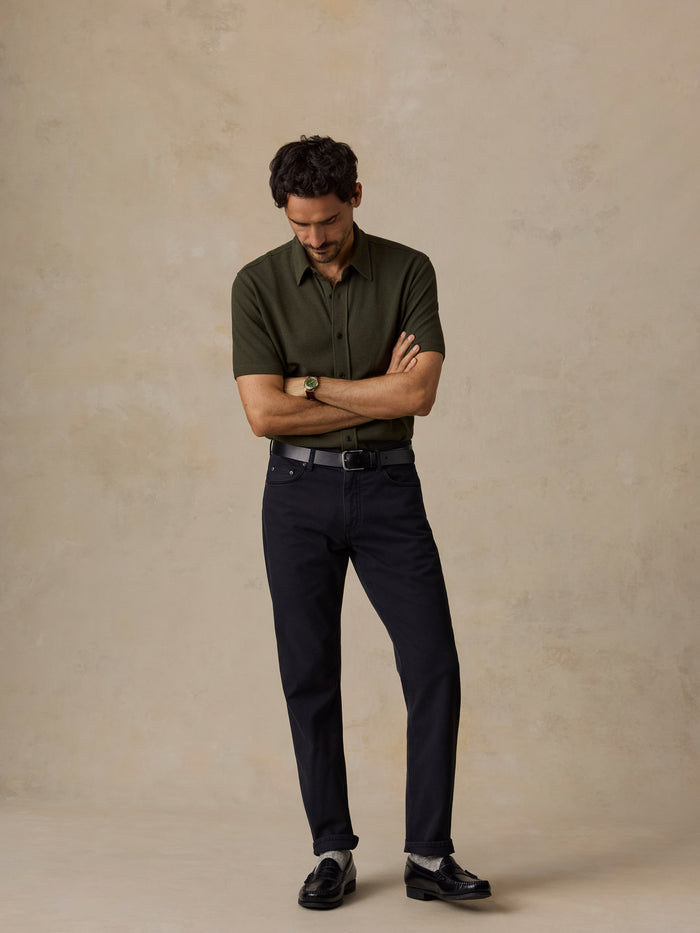 A man stands with arms crossed wearing a dark olive green short-sleeve button-up shirt, dark navy trousers, a black belt, and black loafers against a textured beige studio backdrop.