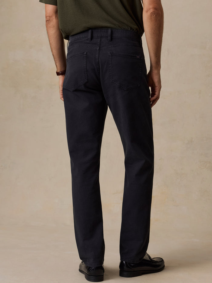 Rear view of a man wearing dark navy straight-leg trousers, a dark olive green t-shirt, and black leather shoes, standing against a textured beige studio backdrop.
