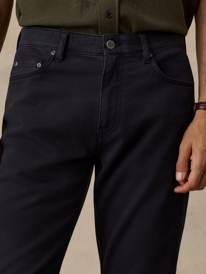 Close-up of a person wearing dark navy blue jeans with a button closure and silver rivets, paired with an olive green shirt, against a neutral beige background.