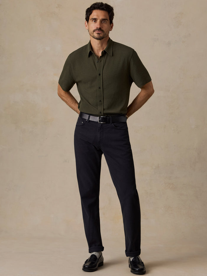 A man modeling a short-sleeve olive green button-down shirt and dark navy trousers, standing against a textured beige studio backdrop.