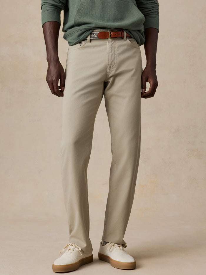 A model wearing a pair of light beige chinos with a brown leather belt, styled with a sage green long-sleeve shirt and off-white sneakers, set against a textured beige studio backdrop.