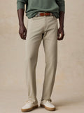 A model wearing a pair of light beige chinos with a brown leather belt, styled with a sage green long-sleeve shirt and off-white sneakers, set against a textured beige studio backdrop.