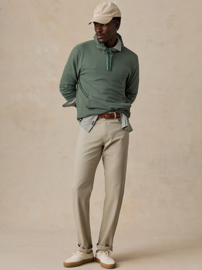 A man modeling a sage green long-sleeve knit polo sweater with a tie-neck detail, styled with beige trousers, a light blue collared shirt, a brown leather belt, and a cream baseball cap against a textured beige studio backdrop.