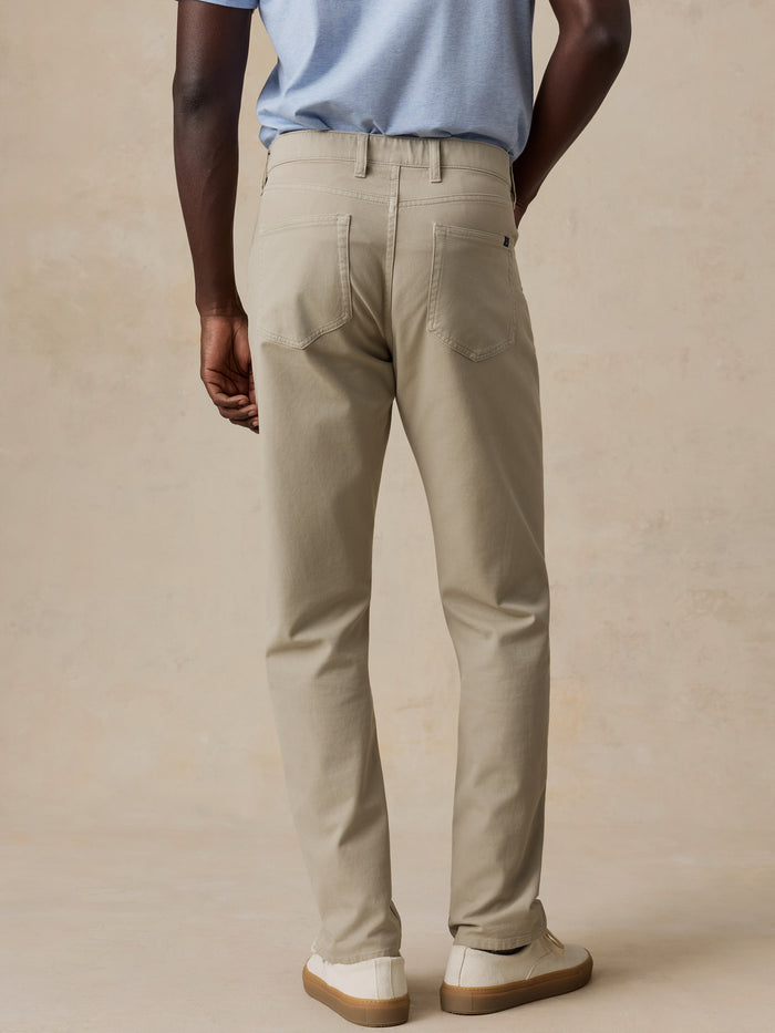 A man shown from the back wearing light beige chino pants, a light blue t-shirt, and white slip-on shoes, standing against a textured beige studio backdrop.